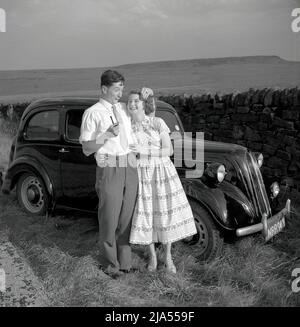 1950s, historisch, ein junges erwachsenes Paar, Freund und Freundin eines Jungen, die an ihrem Wagen der damaligen Zeit, einem Ford Anglia, in einem Grasstreifen an einer Steinmauer, England, Großbritannien, stehen. Dicht beieinander lacht die junge Dame in einem langen Sommerkleid über ihren Freund oder vielleicht Verlobten, der ein lustiges Gesicht macht und eine Pfeife hält. Stockfoto