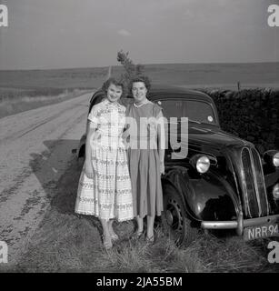 1950s, historisch, zwei Damen, vielleicht Mutter und Tochter, in den langen Kleidern der Ära, die für ihr Foto von ihrem Ford Anglia Auto stehen, das auf einem Grasrand neben einer Steinmauer an einer Landstraße, England, Großbritannien, geparkt ist. Stockfoto