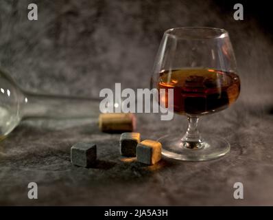 Transparenter Whiskey mit einer Flasche und einem Holzkorken auf dunklem Hintergrund. Whiskey-Steine im Glas Stockfoto