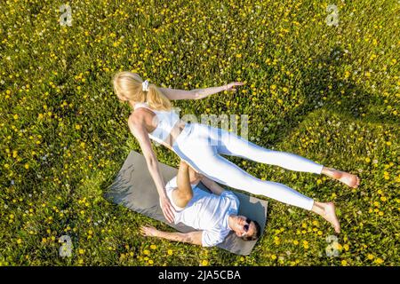 Schönes junges Paar, das im Park Acro Yoga macht. Mann liegt auf Gras und balancierende Frau in den Füßen. Stockfoto
