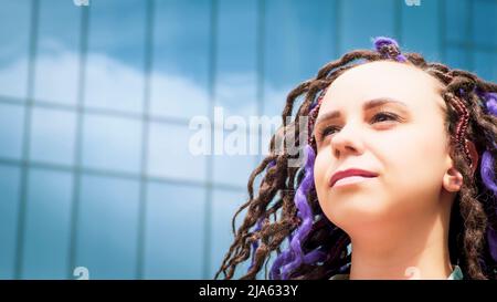 Porträt einer Frau mit Dreadlocks vor dem Hintergrund eines Glasgebäudes. Junge Hündin mit lockiger Frisur, die bei sonnigem Wetter auf verschwommenem Rücken wegschaut Stockfoto