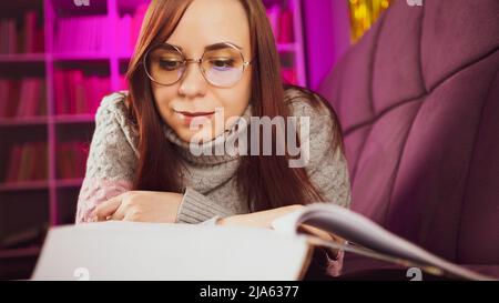 Eine schöne Schülerin mit Brille, die ein großes Buch liest. Eine nachdenkliche junge Frau in einem Pullover, die mit einem Buch auf dem Sofa liegt und abends liest Stockfoto