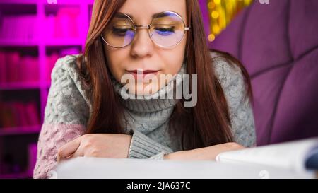 Eine schöne Schülerin mit Brille, die ein großes Buch liest. Eine nachdenkliche junge Frau in einem Pullover, die mit einem Buch auf dem Sofa liegt und abends liest Stockfoto