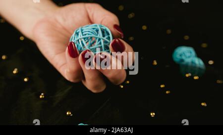 Die Hand einer reifen Frau mit rot polierten Nägeln hält einen geflochtenen Ball auf schwarzem Hintergrund. Nicht erkennbare Person mit Maniküre beschneiden. Konzept der Pflege und Bea Stockfoto