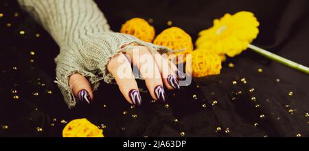 Erwachsene Frauenhand in stylischem Strickhandschuh mit lila Maniküre auf schwarzem Hintergrund. Schneiden Sie nicht erkennbare Person mit Design auf den Nägeln. Konzept der Pflege Stockfoto