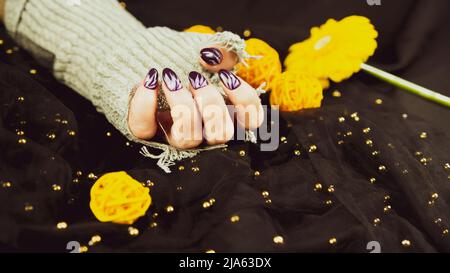 Erwachsene Frauenhand in stylischem Strickhandschuh mit lila Maniküre auf schwarzem Hintergrund. Schneiden Sie nicht erkennbare Person mit Design auf den Nägeln. Konzept der Pflege Stockfoto