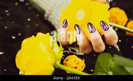 Erwachsene Frauenhand in stylischem Strickhandschuh mit lila Maniküre auf schwarzem Hintergrund. Schneiden Sie nicht erkennbare Person mit Design auf den Nägeln. Konzept der Pflege Stockfoto