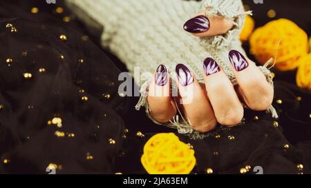 Erwachsene Frauenhand in stylischem Strickhandschuh mit lila Maniküre auf schwarzem Hintergrund. Schneiden Sie nicht erkennbare Person mit Design auf den Nägeln. Konzept der Pflege Stockfoto