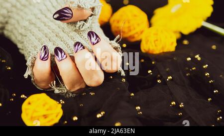 Erwachsene Frauenhand in stylischem Strickhandschuh mit lila Maniküre auf schwarzem Hintergrund. Schneiden Sie nicht erkennbare Person mit Design auf den Nägeln. Konzept der Pflege Stockfoto