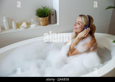 Junge glückliche blonde Frau, die mit Schaum baden und Musik über Kopfhörer hören. Stockfoto
