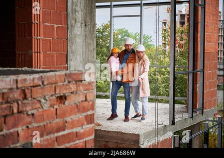 Voller fröhlicher Mann, der seine Tochter hält und lächelt, während er neben seiner Frau auf der Baustelle steht. Glückliche Eltern mit Kind posieren in Wohngebäude im Bau. Stockfoto