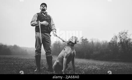 Hunter Mann mit Hund in traditionellen Schießkleidung auf dem Feld halten Schrotflinte, schwarz-weiß Foto. Stockfoto