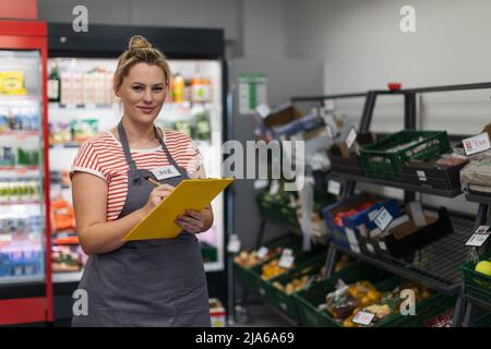 Eine junge Verkäuferin im Supermarkt steht und schreibt vor der Gemüseabteilung. Stockfoto