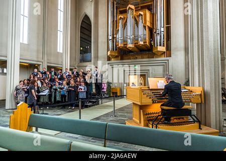 Der Organist Hörður Áskelsson sitzt hier an der Konsole der Hallgrímskirkja in Reykjavík, während ein Chor im Kirchenschiff singt. Der Kirchenmusiker war von 1982 bis 2021 Organist und Kantor der Kirche, gründete 1982 den Hallgrímskirkja Motettenchor und 1996 die Schola Cantorum Reykjavicensis. Die im Hintergrund sichtbare Orgel, gebaut vom Bonner Orgelbauer Johannes Klais, wurde 1992 eingeweiht und hat 72 Register, 102 Reihen und 5.275 Pfeifen. Stockfoto