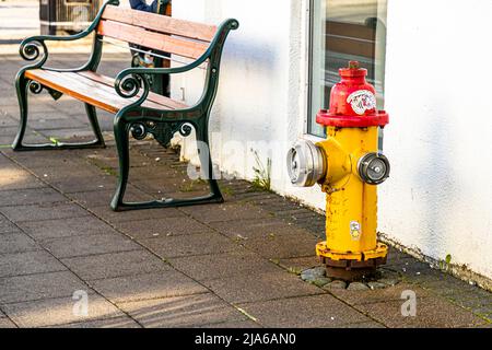 Ein gelb-roter Feuerhydrant steht hier auf einem Gehweg in Reykjavík, Island, neben einer grün gestrichenen Holzbank vor einer weißen Hauswand. Stockfoto