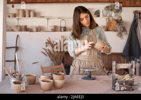 Die junge Kunsthandwerkerin konzentrierte sich während eines Töpferunterrichtete oder Workshops in einem kreativen Studio auf die Bildhauerei einer rohen Tonschale. Frau Keramik Geschäftseigentümer machen Stockfoto