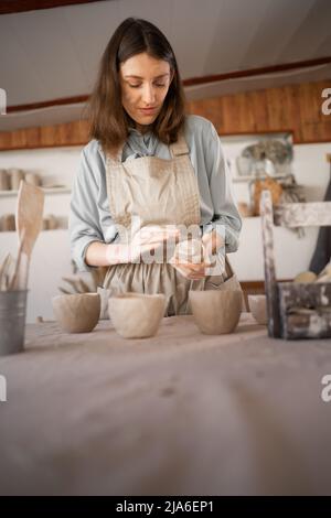 Weibliche Kunsthandwerkerin konzentrierte sich während des Töpferunterrichts oder Workshops im Kreativstudio auf die Modellierung von Krug aus rohem Ton. Frau Keramik Geschäftseigentümer machen Stockfoto