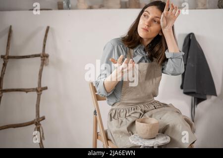Müde kaukasische schöne weibliche Töpfer am Arbeitsplatz. Erstellung von handgefertigten Töpferwaren. Ein Töpfer in einem Keramikstudio bildet einen Topf aus nassem Ton. Damen Stockfoto