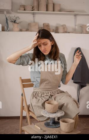 Müde kaukasische schöne weibliche Töpfer am Arbeitsplatz. Erstellung von handgefertigten Töpferwaren. Ein Töpfer in einem Keramikstudio bildet einen Topf aus nassem Ton. Damen Stockfoto