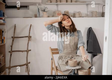 Müde kaukasische schöne weibliche Töpfer am Arbeitsplatz. Erstellung von handgefertigten Töpferwaren. Ein Töpfer in einem Keramikstudio bildet einen Topf aus nassem Ton. Damen Stockfoto