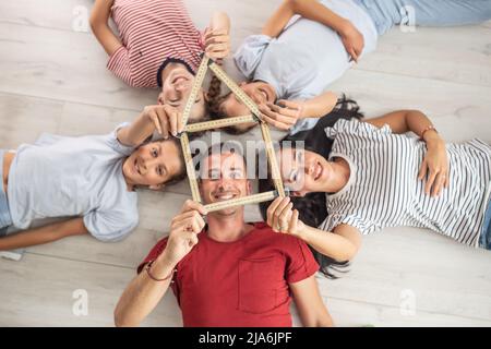 Familie, die auf dem Rücken auf dem Boden liegt, hält eine Silhoutte eines Hauses aus Holzfalzmesser zusammen. Stockfoto