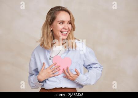 Horizontales mittleres Porträt einer glücklichen, schönen jungen Frau, die ein lässiges Outfit trägt und das pinke Herz aus Papier hält und lächelt Stockfoto