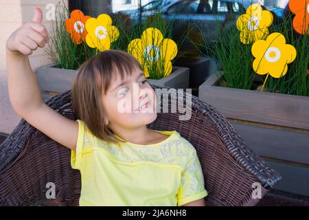 Das glückliche Mädchen sitzt in einem Korbsessel mit der erhobenen Hand. Stockfoto