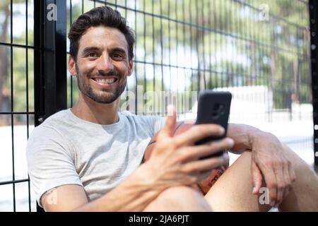 Männlicher Basketballspieler, der sich ausruhen und ein Smartphone benutzen kann Stockfoto