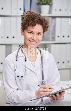 Vertikales mittleres Porträt einer fröhlichen reifen kaukasischen Frau mit lockigen kurzen Haaren, die einen weißen Mantel trägt und als Hausarzt im Krankenhaus arbeitet Stockfoto