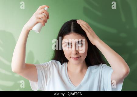 Eine junge schöne Frau sprüht Pflegekosmetik auf ihr Haar. Grüner Hintergrund mit einem Schatten von einem Monsterblatt. Stockfoto