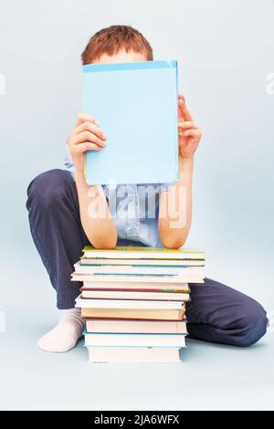 Der Schuljunge sitzt mit einem Stapel Bücher und liest und bedeckt sein Gesicht mit einem isolierten Buch auf blauem Hintergrund Stockfoto