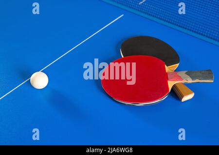 Zwei Schläger für Tischtennis und ein weißer Ball auf dem blauen Tisch. Ein Ping-Pong-Spiel. Sportspiele. Stockfoto