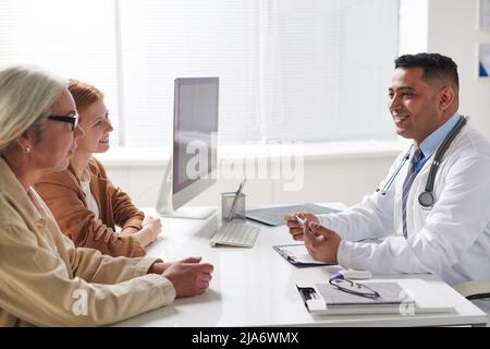 Horizontale Seitenansicht der glücklichen medizinischen Fachärztin aus dem Nahen Osten, die älteren Frauen und ihrer Tochter gute Nachrichten erzählte Stockfoto