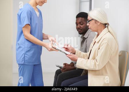 Seitenansicht einer stilvollen älteren Frau, die im Krankenhaus in der Warteschlange sitzt und wichtige Dokumente für medizinische Dokumente unterzeichnet Stockfoto