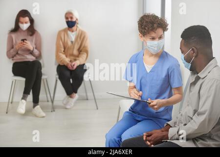 Horizontale Aufnahme eines nicht erkennbaren Mediziners mit Maske, der auf einem Stuhl im Flur neben dem Patienten sitzt und ihm Fragen stellt und Notizen macht Stockfoto