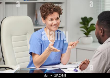 Porträt einer erfolgreichen, reifen kaukasischen Fachärztin, die vor einem nicht erkennbaren Patienten am Tisch sitzt und den Behandlungsplan diskutiert Stockfoto