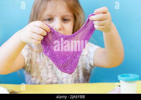 Blonde Mädchen halten und dehnen lila Glitzer Schleim isoliert auf einem blauen Hintergrund. Kind spielt mit einem Schleim Spielzeug. Schleim machen. Speicherplatz kopieren. DIY Konz Stockfoto