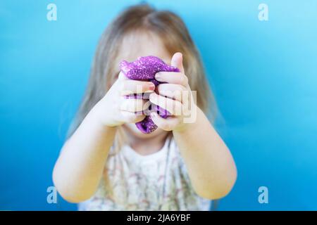 Blonde Mädchen halten und dehnen lila Glitzer Schleim isoliert auf einem blauen Hintergrund. Kind spielt mit einem Schleim Spielzeug. Schleim machen. Speicherplatz kopieren. DIY Konz Stockfoto