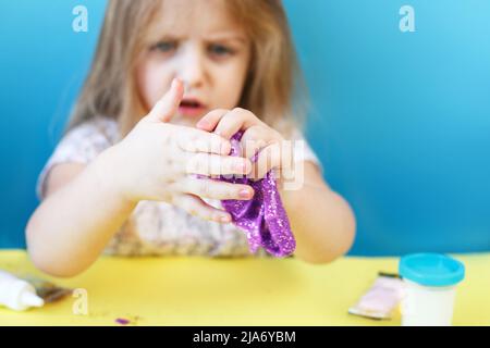 Blonde Mädchen halten lila Glitzer Schleim isoliert auf einem blauen Hintergrund. Kind spielt mit einem Schleim Spielzeug. Schleim hat in den Händen stecken und Kind holen es ab. M Stockfoto