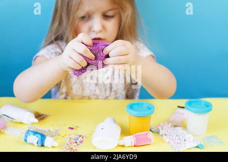 Blonde Mädchen halten lila Glitzer Schleim isoliert auf einem blauen Hintergrund. Kind spielt mit einem Schleim Spielzeug. Schleim machen. Speicherplatz kopieren. DIY-Konzept. Stockfoto