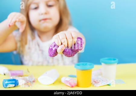 Blonde Mädchen halten lila Glitzer Schleim isoliert auf einem blauen Hintergrund. Kind spielt mit einem Schleim Spielzeug. Schleim machen. Speicherplatz kopieren. DIY-Konzept. Stockfoto