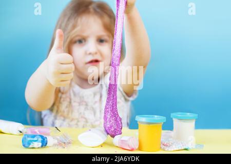 Blonde Mädchen halten lila Glitzer Schleim und machen Daumen nach oben Zeichen isoliert auf einem blauen Hintergrund. Kind spielt mit einem Schleim Spielzeug. Schleim machen. Speicherplatz kopieren. Stockfoto