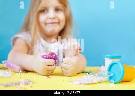 Blonde Mädchen halten lila Glitzer Schleim und machen Daumen nach oben Zeichen isoliert auf einem blauen Hintergrund. Kind spielt mit einem Schleim Spielzeug. Schleim machen. Speicherplatz kopieren. Stockfoto