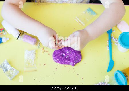 Schleim zu Hause machen. DIY Concept.Kids Hände machen lila Schleim mit Pailletten auf einem gelben Hintergrund. Flach liegend, Draufsicht. Stockfoto