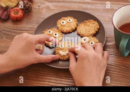 Hände, die Pudergesicht auf Zuckerkeks machen, die auf den Teller gelegt werden, um Süßigkeiten für Halloween vorzubereiten Stockfoto