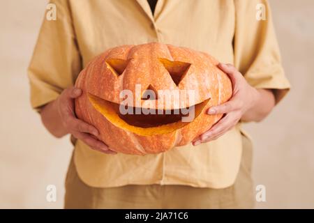 Nahaufnahme einer nicht erkennbaren Frau im gelben Outfit, die einen witzig geschnitzten Kürbis hält, während sie sich auf Halloween vorbereitete Stockfoto