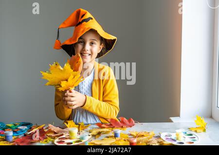 Kaukasisches Mädchen in einem Hexenkostüm sitzt zu Hause am Tisch und bereitet Kunsthandwerk für den Halloween-Urlaub vor. DIY und Dekor im Zimmer gemacht Stockfoto