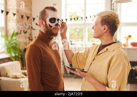 Lächelnde junge blonde Frau in gelbem Hemd, der den Mann schminkt, während sie ihn auf die Halloween-Party vorbereitet Stockfoto
