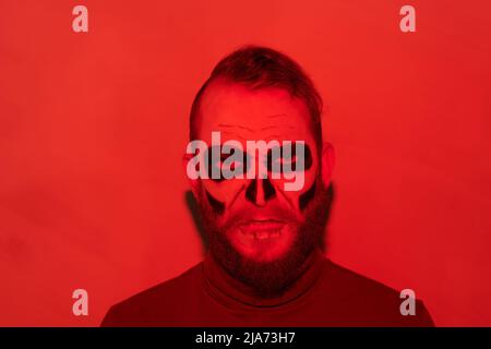 Porträt eines wütenden bärtigen Zombie-Mannes mit düsterem Make-up im roten Licht stehend, Halloween-Konzept Stockfoto