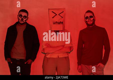 Gruppe von jungen Freunden mit erschreckenden Make-ups und Person mit unzufriedenem Gesicht auf Papiertüte stehend in Reihe, Rotlichteffekt Stockfoto